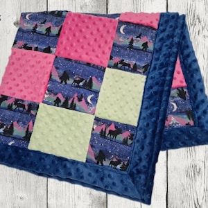 May include: A patchwork blanket with a blue minky border. The blanket features squares of pink, light green, and a patterned fabric with a night sky, mountains, and Bigfoot silhouettes. The blanket is on a white wood surface.
