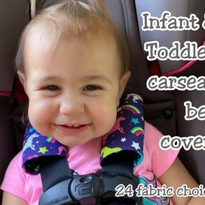 May include: A smiling toddler wearing car seat belt covers. The covers are patterned with unicorns, rainbows, and stars on a dark blue background. The text on the image reads "Infant & Toddler carseat belt covers" and "24 fabric choices."
