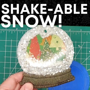 May include: A clear glass snow globe ornament with a glittery white snow effect. The globe contains a colorful paper design with a star and swirls. The base of the globe is brown and has a metal loop for hanging.
