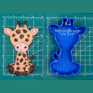 May include: A blue silicone mold of a baby giraffe, sitting with its legs crossed. The mold is on a green cutting mat with inch markings. The text "Baby Giraffe, Large ~3.25" x 5.5"" is printed on the mold.