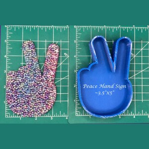 May include: A blue silicone mould of a peace sign hand gesture. The mould is on a green cutting mat with white markings. The mould is approximately 3.5 inches by 5 inches. The text "Peace Hand Sign ~3.5" x 5" is printed on the mould.
