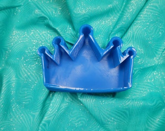 May include: A blue plastic crown-shaped mold for baking or crafting. The mold is sitting on a turquoise fabric with a sparkly texture.