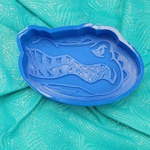 May include: A blue alligator-shaped tray with detailed features, including teeth and scales. The tray is made of a solid blue material and is set against a teal fabric background.