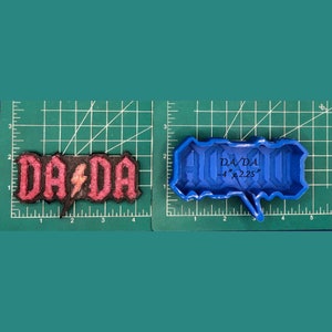 May include: A blue silicone mould for baking a cake or cookie in the shape of the words "DA DA" with a lightning bolt in between. The mould is 4 inches wide and 2.25 inches tall.