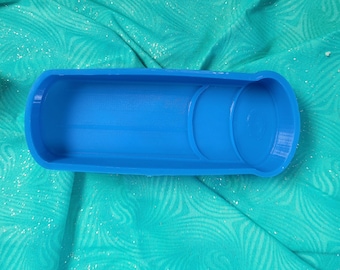 Puede incluir: Un molde de plástico azul para hacer jabón o velas. El molde tiene una forma rectangular con un borde redondeado y una pequeña hendidura circular en un lado.