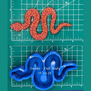 Puede incluir: Un molde de silicona azul con forma de serpiente. El molde está sobre una estera de corte verde con marcas de pulgada blancas. El texto "Snake - Full Body ~6"x3.25"" está impreso en el molde.