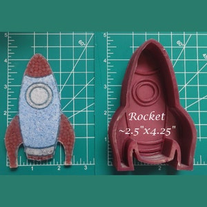 May include: A red silicone mold of a rocket ship with the text "Rocket ~2.5"x4.25"" on the mold. The mold is on a green grid background.