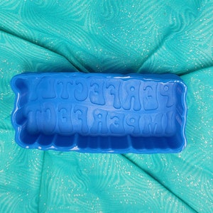 May include: Blue silicone mould for making soap or candles. The mould is shaped like a rectangle and has the words "Imperfectly Perfect" written in raised letters.