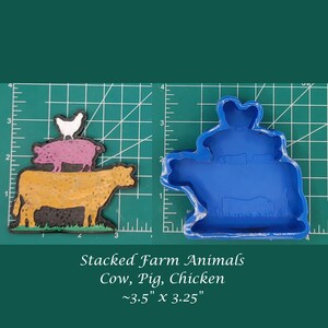 May include: A blue silicone mold for baking cookies in the shape of a cow, pig, and chicken stacked on top of each other. The mold is on a green cutting mat with white markings. The text "Stacked Farm Animals Cow, Pig, Chicken ~3.5" x 3.25"" is below the mold.