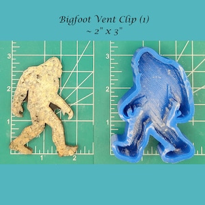 Puede incluir: Un recorte de madera de Bigfoot y un molde de silicona azul de Bigfoot, ambos de aproximadamente 5 cm por 7,5 cm, se muestran en una estera de corte verde con líneas de cuadrícula blancas.