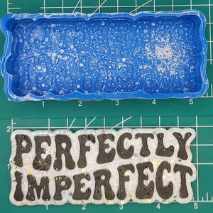 Puede incluir: Un molde de silicona azul con el texto "Perfectly Imperfect" en letras en relieve. El molde está sobre una estera de corte verde. Debajo del molde hay una pieza de resina blanca con el texto "Perfectly Imperfect" en letras negras.
