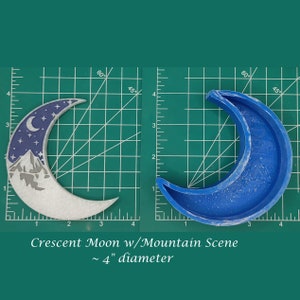 May include: Two crescent moon-shaped objects. One is a finished product with a mountain scene and starry sky design, approximately 4 inches in diameter. The other is a blue mold of the same shape. The text reads "Crescent Moon w/Mountain Scene ~ 4" diameter."