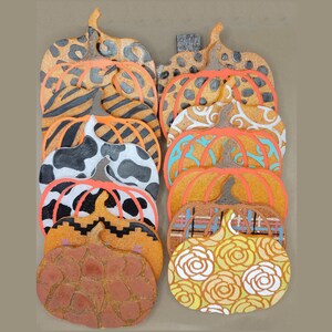 May include: A stack of 12 pumpkin-shaped coasters with various patterns, including animal print, plaid, and floral designs. The coasters are made of cork and have a natural brown colour.