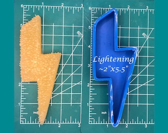 May include: Blue plastic lightning bolt cookie cutter on a green cutting mat. The cutter measures approximately 2 inches by 5.5 inches.