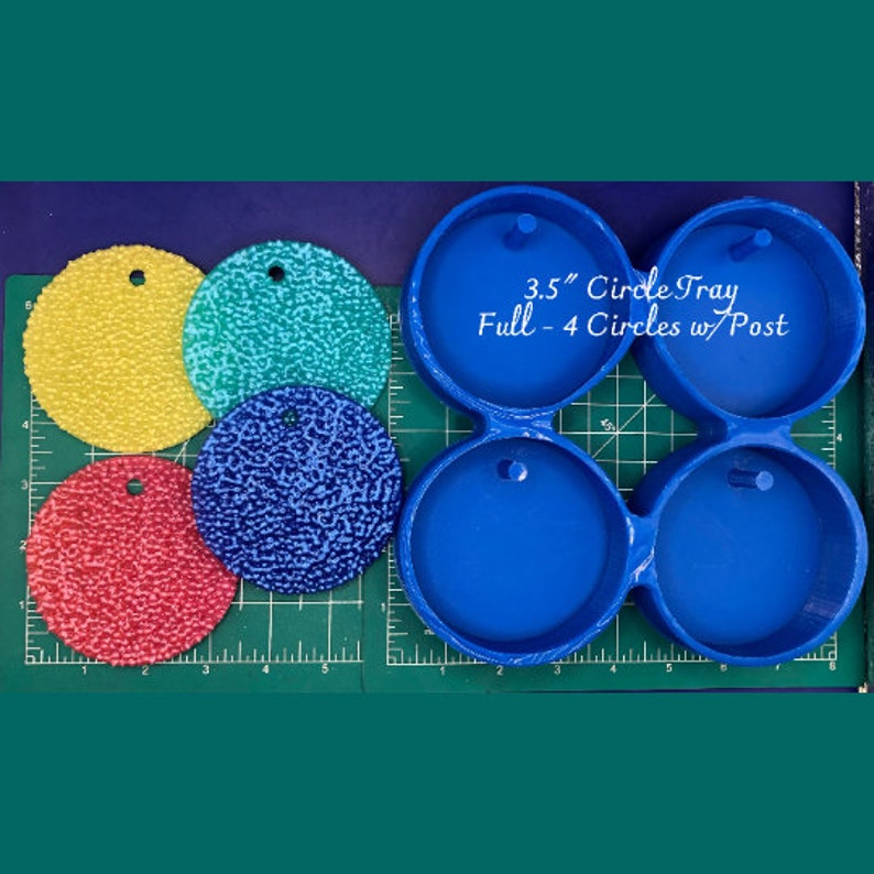 Op de afbeelding: Blauwe kunststof mal voor het maken van 3,5 inch hars cirkels. De mal heeft vier ronde holtes en een pen in het midden. De mal wordt getoond met vier afgewerkte hars cirkels in geel, groen, blauw en rood.