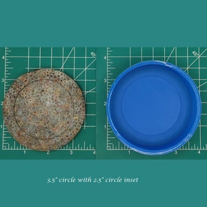 Op de afbeelding: Een cirkel met een diameter van 8,9 cm met een ingelegde cirkel met een diameter van 6,4 cm. De buitenste cirkel is gemaakt van een bruin materiaal met een textuur. De binnenste cirkel is gemaakt van blauw siliconen.