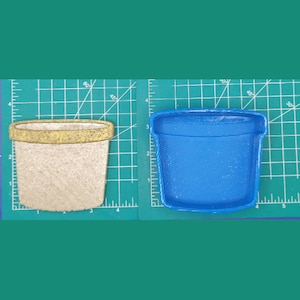 May include: Two cookie cutters in the shape of a flower pot. One is a light beige color with a gold rim, and the other is blue. The cookie cutters are on a green cutting mat.