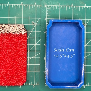 May include: A red soda can with a textured surface and a white top, next to a blue mold. The mold is labeled "Soda Can ~2.5"x4.5"" on a green cutting mat.