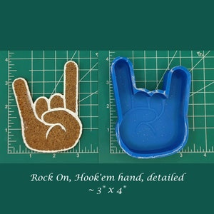 May include: A blue silicone mold of a hand making the rock on gesture. The mold is on a green cutting mat with white markings. The mold is approximately 3 inches by 4 inches. "Rock On, Hook'em hand, detailed ~ 3" x 4" "