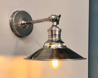 May include: A silver-toned wall-mounted light fixture with a conical shade and a visible lightbulb. The fixture has an adjustable arm and a circular base. The light casts a warm glow on the wall.