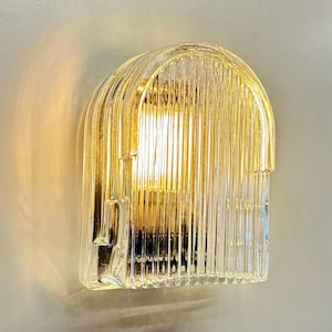 May include: A clear glass wall sconce with a ribbed, arched design. The light fixture has a warm, yellow glow emanating from within. The sconce is mounted on a light gray wall, creating a modern aesthetic.