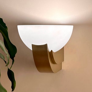 May include: A gold wall sconce with a white frosted glass shade. The sconce has a curved design and is mounted on a wall.