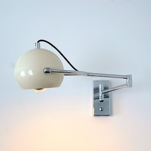 Puede incluir: Una lámpara de pared esférica blanca con un brazo y una base cromados. La lámpara está encendida y proyecta una luz cálida.