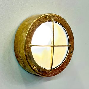 Puede incluir: Aplique de pared redondo de color latón con diseño de ojo de buey. La lámpara tiene una lente de vidrio transparente con una rejilla metálica en forma de cruz. El aplique está montado en una pared gris claro.