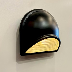 Könnte beinhalten: Eine schwarze, kuppelförmige Wandleuchte mit einem strukturierten, gelb getönten Lichtpaneel. Die Leuchte ist an einer hellgrauen Wand montiert. Die Leuchte hat ein modernes, minimalistisches Design.