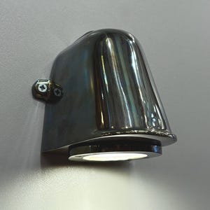 May include: A chrome-finished, wall-mounted light fixture. The fixture has a rounded, tapered shape with a circular light source at the bottom. Two screws secure a small, circular mounting plate to the wall. The light is illuminated.