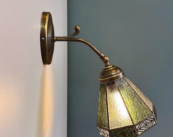 May include: A wall-mounted brass sconce with a green, geometric-patterned glass shade. The fixture has a curved arm and a circular base. The shade is a light green color and the metal has a vintage finish.