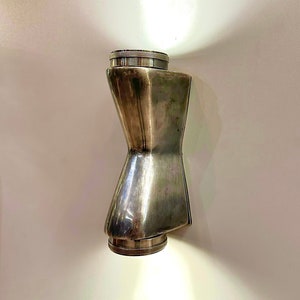 Puede incluir: Una lámpara de pared de metal plateado con una forma de reloj de arena única. La lámpara tiene dos bombillas que emiten una luz cálida.