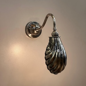 May include: A silver wall sconce featuring a curved arm and a teardrop-shaped shade with a textured, ridged design. The sconce is mounted on a circular base. The fixture is made of metal.