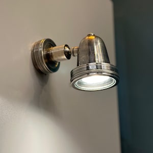 Puede incluir: Un foco plateado montado en la pared con una base circular y una cabeza en forma de campana. La luz está encendida, iluminando el área. El accesorio tiene un diseño decorativo y estriado alrededor de la base y la abertura de la luz.
