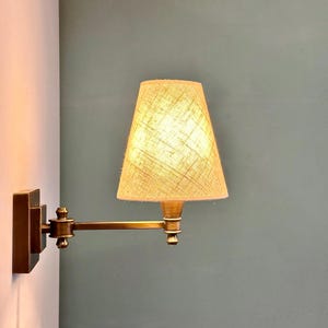 K&ouml;nnte beinhalten: Eine Wandleuchte mit Messing-Finish. Die Lampe hat einen rechteckigen Sockel, einen verstellbaren Arm und einen beigefarbenen Lampenschirm. Der Lampenschirm hat ein strukturiertes Aussehen, und das Licht ist an und beleuchtet den Schirm.