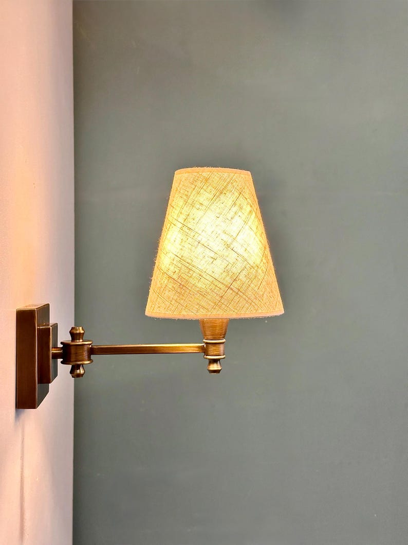 K&ouml;nnte beinhalten: Eine Wandleuchte mit Messing-Finish. Die Lampe hat einen rechteckigen Sockel, einen verstellbaren Arm und einen beigefarbenen Lampenschirm. Der Lampenschirm hat ein strukturiertes Aussehen, und das Licht ist an und beleuchtet den Schirm.