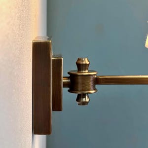 K&ouml;nnte beinhalten: Nahaufnahme einer bronzefarbenen Wandleuchte. Die Leuchte hat eine rechteckige Basis und einen schwenkbaren Arm. Das Design weist ein dekoratives, detailliertes Gelenk und ein kleines, zylindrisches Element auf. Der Hintergrund ist eine hellbeige Wand und eine t&uuml;rkisblaue Wand.