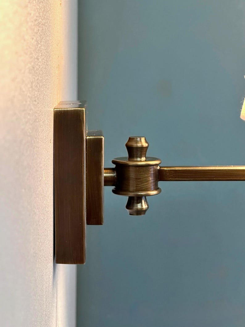 K&ouml;nnte beinhalten: Nahaufnahme einer bronzefarbenen Wandleuchte. Die Leuchte hat eine rechteckige Basis und einen schwenkbaren Arm. Das Design weist ein dekoratives, detailliertes Gelenk und ein kleines, zylindrisches Element auf. Der Hintergrund ist eine hellbeige Wand und eine t&uuml;rkisblaue Wand.