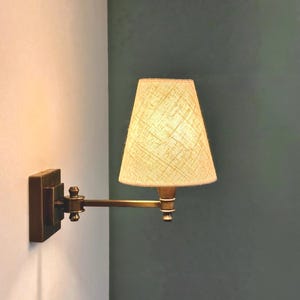 K&ouml;nnte beinhalten: Eine Wandleuchte mit rechteckigem Sockel und verstellbarem Arm. Die Lampe hat einen beigefarbenen Stoffschirm und eine Messingoberfl&auml;che. Sie ist an einer wei&szlig;en Wand mit einer dunkelgr&uuml;nen Wand im Hintergrund befestigt.