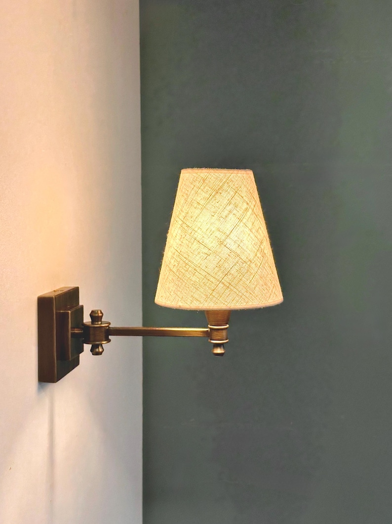 K&ouml;nnte beinhalten: Eine Wandleuchte mit rechteckigem Sockel und verstellbarem Arm. Die Lampe hat einen beigefarbenen Stoffschirm und eine Messingoberfl&auml;che. Sie ist an einer wei&szlig;en Wand mit einer dunkelgr&uuml;nen Wand im Hintergrund befestigt.