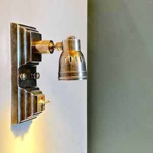 May include: A vintage-style brass wall-mounted light fixture with a round, metal shade and a toggle switch. The light is mounted on a white wall.
