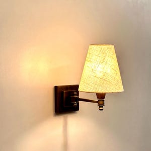 K&ouml;nnte beinhalten: Eine Wandleuchte mit quadratischem, dunkelbronzenem Sockel und beigefarbenem Stoffschirm. Die Lampe hat einen verstellbaren Arm und ein warmes, einladendes Licht, perfekt f&uuml;r die Ambientebeleuchtung in einem Schlaf- oder Wohnzimmer.