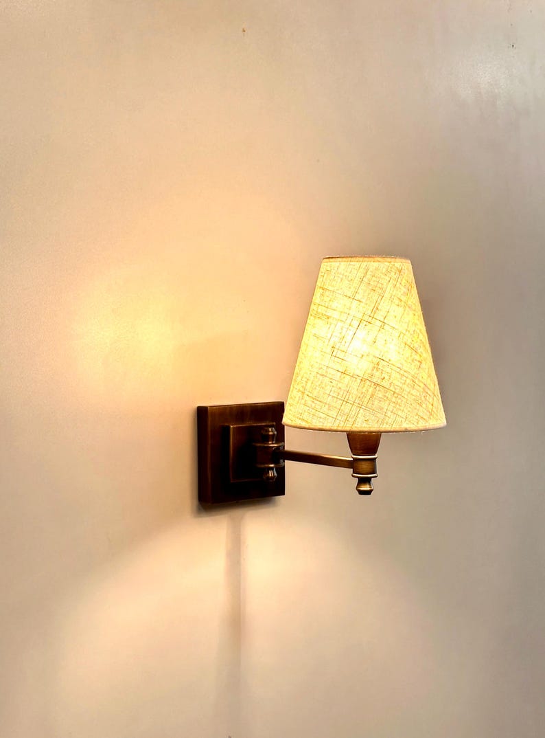 K&ouml;nnte beinhalten: Eine Wandleuchte mit quadratischem, dunkelbronzenem Sockel und beigefarbenem Stoffschirm. Die Lampe hat einen verstellbaren Arm und ein warmes, einladendes Licht, perfekt f&uuml;r die Ambientebeleuchtung in einem Schlaf- oder Wohnzimmer.