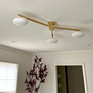 Puede incluir: Una moderna lámpara de techo con acabado dorado y tres pantallas de globo de vidrio esmerilado. La lámpara está montada en un techo blanco y tiene un diseño minimalista. La lámpara es una adición elegante a cualquier habitación.