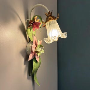 May include: A wall-mounted light fixture with a floral design. The fixture features a white curved arm, a frosted glass bell-shaped shade, and decorative flowers in pink, green, and brown. The light is on, casting a shadow on the wall.