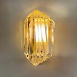 Puede incluir: Aplique de pared de vidrio transparente con un diseño acanalado y un brillo amarillo cálido. La lámpara tiene una forma geométrica y está montada en una pared gris. El aplique es un elemento de iluminación decorativo.