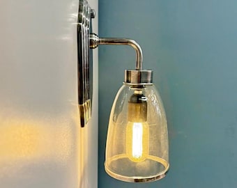 May include: A wall-mounted light fitting with a clear glass shade and a silver-toned metal frame. The light bulb is illuminated, casting a warm glow. The fitting is mounted on a white and blue wall.