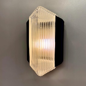May include: A modern wall sconce with a black frame and a clear, ribbed glass front. The light fixture has a geometric shape and emits a warm glow. The sconce is mounted on a gray wall.