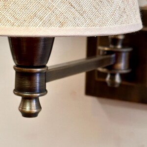 K&ouml;nnte beinhalten: Nahaufnahme einer Wandleuchte mit Bronze-Finish. Die Lampe hat einen beigefarbenen Leinen-Lampenschirm und einen verstellbaren Arm. Die Leuchte ist mit einer klassischen, Vintage-&Auml;sthetik gestaltet, geeignet f&uuml;r die Innenbeleuchtung.