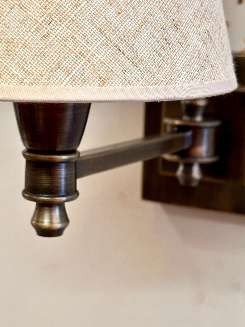 K&ouml;nnte beinhalten: Nahaufnahme einer Wandleuchte mit Bronze-Finish. Die Lampe hat einen beigefarbenen Leinen-Lampenschirm und einen verstellbaren Arm. Die Leuchte ist mit einer klassischen, Vintage-&Auml;sthetik gestaltet, geeignet f&uuml;r die Innenbeleuchtung.